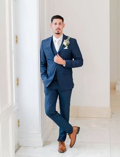 a man in a blue suit standing next to a white wall and looking at the camera