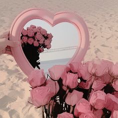 a pink heart shaped mirror sitting on top of a sandy beach next to some flowers