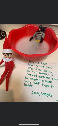 an elf is sitting in front of a red bowl with water on it and a note written to him