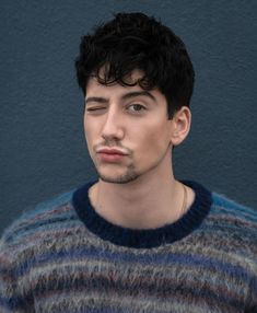 a young man with black hair and a moustache looks at the camera while wearing a striped sweater