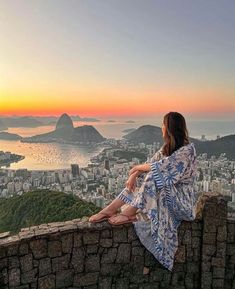 a woman sitting on top of a stone wall looking at the city and mountains in the distance