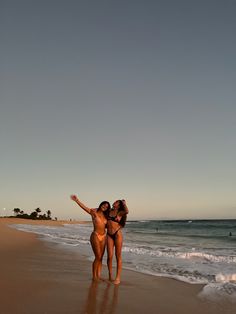 two women in bikinis standing on the beach with their arms around each other,