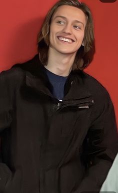 a woman standing in front of a red wall wearing a black jacket and smiling at the camera