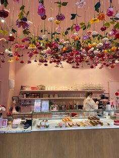 a woman is behind the counter at a pastry shop with flowers hanging from the ceiling