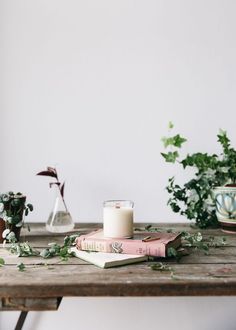 a candle and some books on a table with plants in vases behind the candles