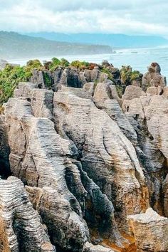 some very pretty rocks by the water with trees on top and mountains in the background