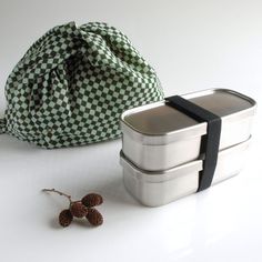 three stainless steel lunch boxes with black straps and two small pine cones on the side