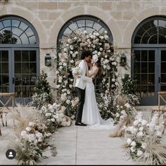 a bride and groom standing in front of a floral arch with white flowers on it