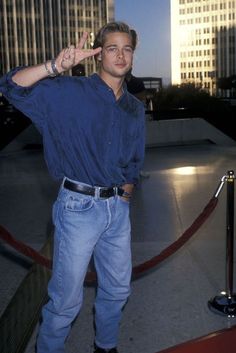 a man standing in front of tall buildings giving the peace sign with his hand while wearing blue jeans and black boots