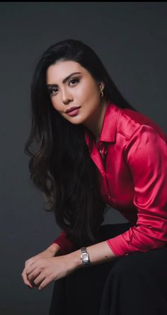 a woman with long black hair wearing a red shirt and black pants sitting on a chair