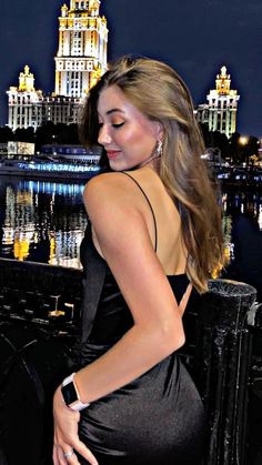 a beautiful woman in a black dress standing next to a body of water at night