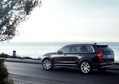 a black volvo suv driving down the road next to the ocean and trees in front of it