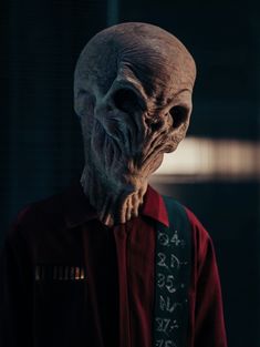 an alien man wearing a red shirt and black vest with writing on it's chest