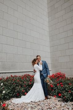 a bride and groom standing in front of some red flowers at their wedding day, posing for the camera