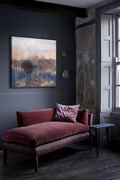 a red couch sitting next to a window on top of a wooden floor in front of a painting