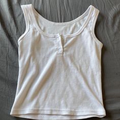 That Tank Top Is A Classic White Color. It's Sleeveless And Looks Really Comfy. The Ribbed Texture Adds A Nice Touch. There's A Single Button Closure In The Front. The Gray Surface Makes The Shirt Stand Out. The White Color Gives A Clean And Fresh Look. The Sleeveless Design Is Perfect For Warm Days. Ribbed Texture Adds A Subtle Detail To The Top. The Button Closure Adds A Stylish Element. It Seems Like A Versatile Piece For Any Outfit. The White Color Is Timeless And Easy To Match. The Ribbed T White Buttoned Tank Top For Summer, Casual Buttoned Tank Top, Casual Sleeveless Crop Top With Button Closure, White Casual Crop Top With Buttons, Ribbed Tank Top Outfit, Casual White Buttoned Tank Top, Casual Cami Tank Top With Buttons, White Tank Top With Buttons, Sleeveless Cotton Crop Top With Buttons