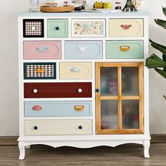 a white dresser with drawers and pictures on the top shelf, next to a potted plant