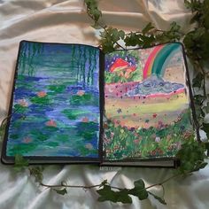 an open book with paintings on it sitting on a bed next to some leaves and flowers