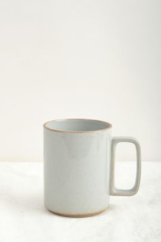 a white coffee mug sitting on top of a white tableclothed surface with an empty cup in the middle