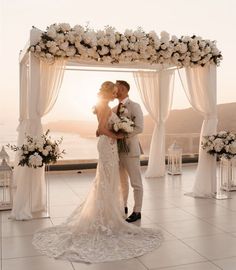 a bride and groom kissing under an arch decorated with white flowers on the beach at sunset