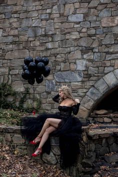 a woman in a black dress sitting on a stone wall with balloons floating above her