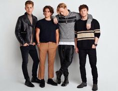 four young men standing next to each other in front of a white wall wearing black pants and sweaters