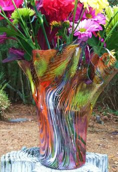 a colorful vase filled with lots of flowers on top of a wooden stump in the grass