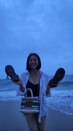 a woman standing on top of a beach next to the ocean holding two pairs of shoes