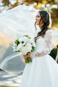 a woman in a wedding dress holding a bouquet and veil over her head while standing outside