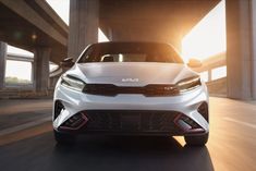 the front end of a white car driving on a road under an overpass at sunset