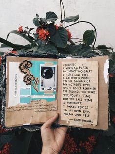 a hand holding an open book in front of some flowers and plants with writing on it