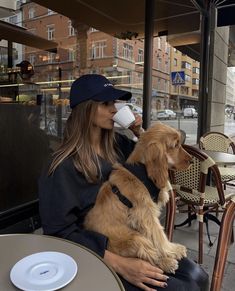 a woman sitting at an outdoor table drinking from a coffee cup while holding her dog