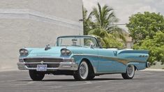 an old blue car parked in a parking lot next to a white wall and palm trees