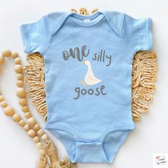 a baby bodysuit with the words, one silly goose on it and tassels next to it