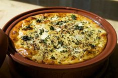 a pizza with cheese and spinach in a clay dish on a table top next to a potted plant