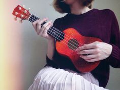 a woman holding a red ukulele in her right hand and wearing a white skirt