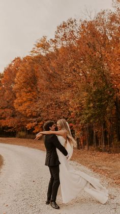 a bride and groom are hugging on the road in front of trees with orange leaves