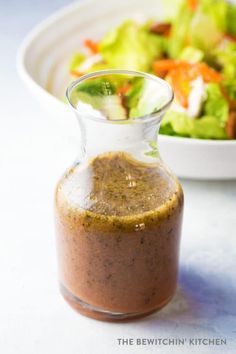 a glass bottle filled with dressing next to a bowl of salad on a white table