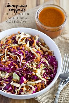 a white bowl filled with coleslaw next to a fork and spoon on top of a wooden table