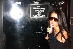 a woman standing in front of a black wall holding her finger up to the camera