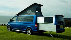 a blue van parked on top of a lush green field next to the ocean with an awning