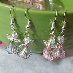 two silver angel earrings with pink crystal stones on them next to a green bowl full of flowers