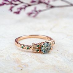 Inspired by nature's delicate beauty, the Moss Blossom ring features a round-cut natural moss agate as the centerpiece, symbolizing the serene connection between earth and art. The band is adorned with petal-like designs on each side, reminiscent of a blooming flower cradling a vibrant green fruit. Each petal is embellished with moissanite, adding a subtle sparkle that enhances the ring’s organic charm. The design seamlessly blends natural inspiration with elegance, perfect for those who appreciate understated sophistication in everyday wear. 𝗗𝗘𝗧𝗔𝗜𝗟𝗦𝗠𝗔𝗜𝗡 𝗦𝗧𝗢𝗡𝗘𝗚𝗲𝗺𝘀𝘁𝗼𝗻𝗲: Natural Moss Agate𝗖𝗼𝗹𝗼𝗿: Green𝗦𝗵𝗮𝗽𝗲: Round 5mm𝗖𝘂𝘁: Full𝗠𝗲𝗮𝘀𝘂𝗿𝗲𝗺𝗲𝗻𝘁: 1.0 ct approx𝗤𝘂𝗮𝗹𝗶𝘁𝘆: VS clarity grademoss agate are natural and can therefore slightly vary in color Nature-inspired May Birthstone Jewelry, Nature-inspired Gemstone Rings For Promise, Nature-inspired Round Flower Ring For Anniversary, Nature-inspired Gemstone Rings For May Birthstone, Nature-inspired Rings With May Birthstone, Nature-inspired May Birthstone Promise Ring, Nature-inspired Wedding Rings With Natural Stones, Nature-inspired Promise Ring With May Birthstone, Elegant Round Opal Ring With Natural Inclusions