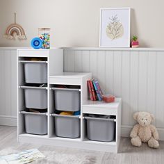 a white bookcase with several bins and a teddy bear sitting on the floor