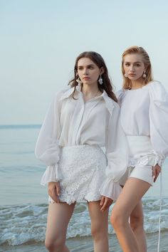 two young women standing next to each other on the beach wearing white clothing and high heels