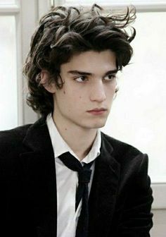 a young man with curly hair wearing a suit and tie looking at the camera while sitting in front of a window