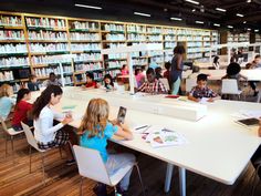 children are sitting at tables in the library and looking at books on shelves behind them