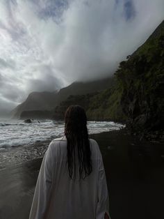 A picture of a girl on the Seixal beach on Madeira. Her wet hair from the sea is falling down onto her white shirt. She is facing the sea, not the camera. The sky is cloudy without the sun brightening it up, so the colours seem dark. Black Sand Beach Photo Ideas, Black Sand Beach Aesthetic, Seixal Beach Madeira, Madeira Outfit Ideas, Black Sand Aesthetic, Black Beach Aesthetic, Cloudy Beach Aesthetic, Madeira Portugal Aesthetic, Wet Hair Aesthetic