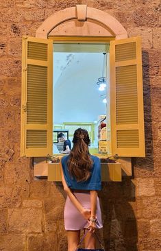 a woman standing in front of a window with yellow shutters and holding a handbag