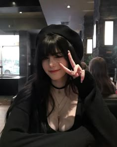 a woman with long black hair wearing a hat and making the peace sign in front of her face